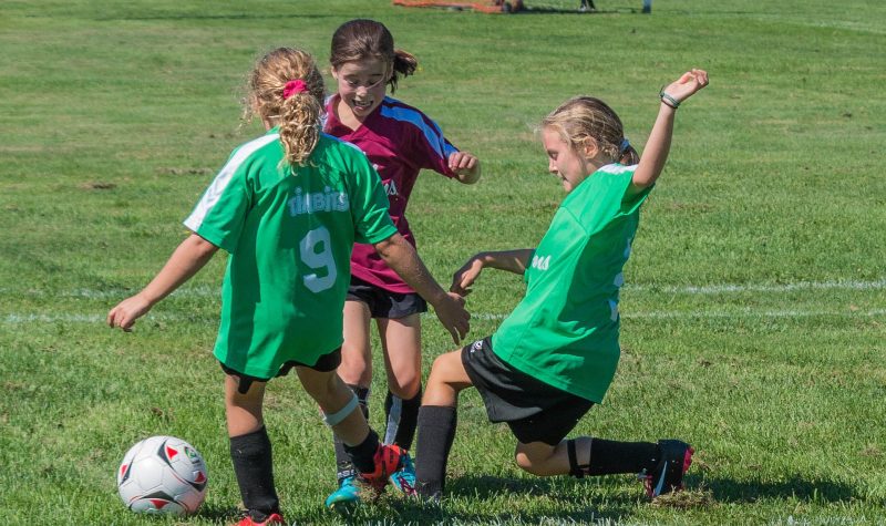 3 jeunes filles jouent au soccer, 2 sont en chandail vert et la troisième est en chandail bordeaux