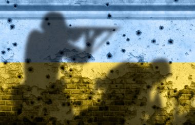 Les couleurs du drapeau de l'Ukraine bleu et jaune sur un mur criblé de balles avec l'ombre de deux soldats armée dessus.