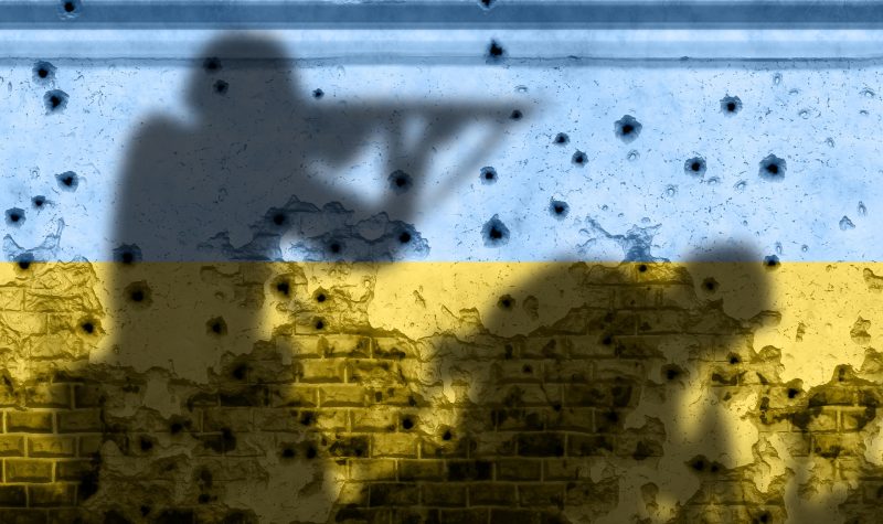 Les couleurs du drapeau de l'Ukraine bleu et jaune sur un mur criblé de balles avec l'ombre de deux soldats armée dessus.