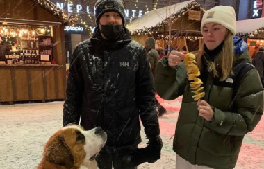 A man and a woman wearing winter clothing stand side-by-side, the woman ripping off a piece of the snack she is holding while a big dog looks up at her enviously.