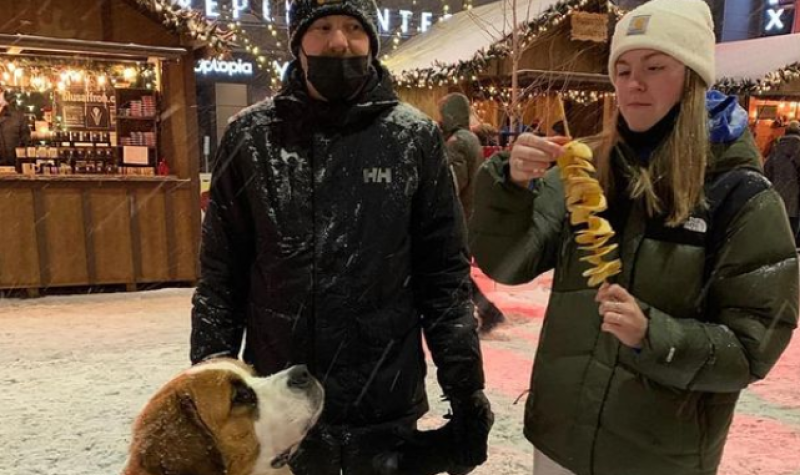 A man and a woman wearing winter clothing stand side-by-side, the woman ripping off a piece of the snack she is holding while a big dog looks up at her enviously.