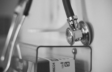 A stethoscope hangs on a white wall and a plastic container hangs underneath it.