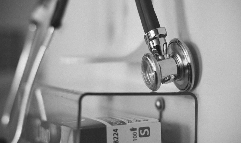 A stethoscope hangs on a white wall and a plastic container hangs underneath it.