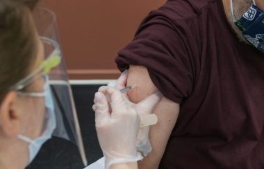 A person with a face shield and medical mask inserts a needle into the arm of a person wearing a brown shirt.