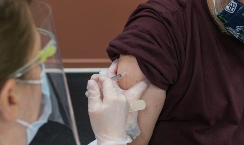 A person with a face shield and medical mask inserts a needle into the arm of a person wearing a brown shirt.