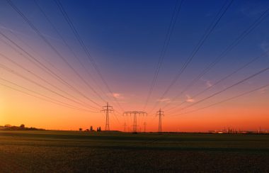 Sunset over a bare field with energy transformers in the middle of them. The wires from the transformers reach across the top of the picture.