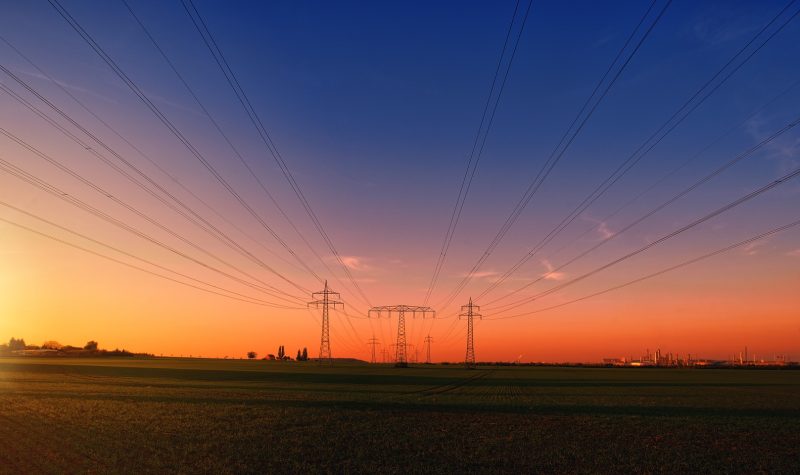 Sunset over a bare field with energy transformers in the middle of them. The wires from the transformers reach across the top of the picture.