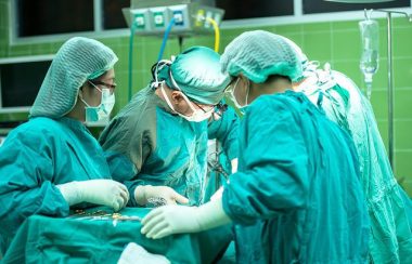 4 chirurgiens autour d'une table d'opération. Les chirurgiens sont habillés d'un sarot bleu-vert. Leurs têtes sont recouvertes de filets bleu-vert. Ils portent tous des gants de chirurgies.
