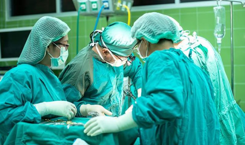 4 chirurgiens autour d'une table d'opération. Les chirurgiens sont habillés d'un sarot bleu-vert. Leurs têtes sont recouvertes de filets bleu-vert. Ils portent tous des gants de chirurgies.