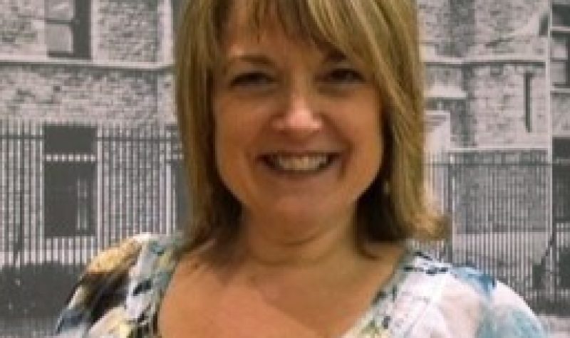 Susan Walsh is smiling in this photo. There is a greyish background and she is wearing a white and coloured blouse.