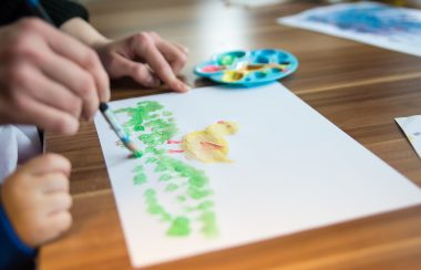 Un enfant fait un dessein au pinceau avec une palette de couleurs. Il représente un poussin dans l'herbe.