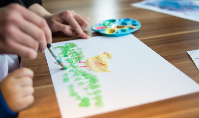 Un enfant fait un dessein au pinceau avec une palette de couleurs. Il représente un poussin dans l'herbe.