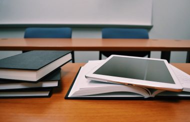 A tablet, and a few books sit stacked on a school desk. In the background a brown school table and 2 blue chairs.