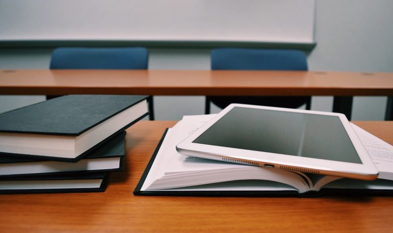 A tablet, and a few books sit stacked on a school desk. In the background a brown school table and 2 blue chairs.