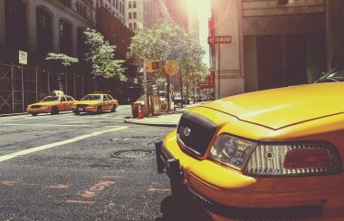 A yellow taxi sits at an intersection with sunlight shining upon it and trees and buildings lining both street sides. At the other side of the intersection 2 more taxis sit at the intersection.