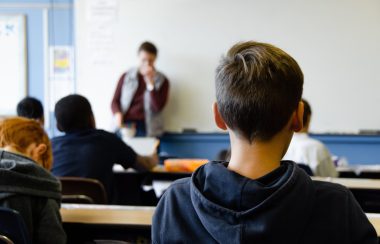 Students in a classroom listen to a teacher during a classroom session in a blue and white room.