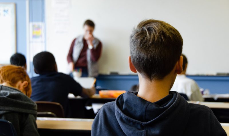 Students in a classroom listen to a teacher during a classroom session in a blue and white room.