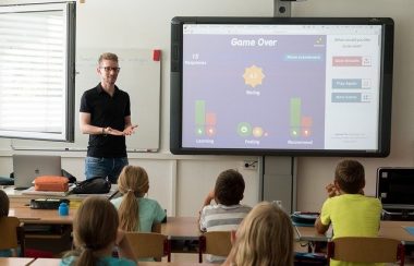 Image d'un enseignant qui enseigne devant la classe. À droite de l'enseignant se trouve un tableau interactif. 5 élèves apparaissent sur l'image, assis à leur place devant leur pupitre.