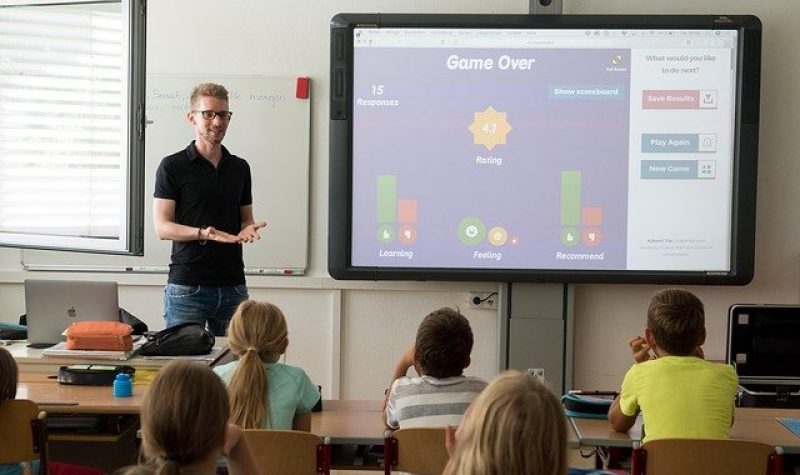 Un professeur qui enseigne devant une classe d'une école primaire. On aperçoit un tableau interactif à l'avant de la classe. 6 élèves sont assis derrière leur pupitre dans les première rangées.