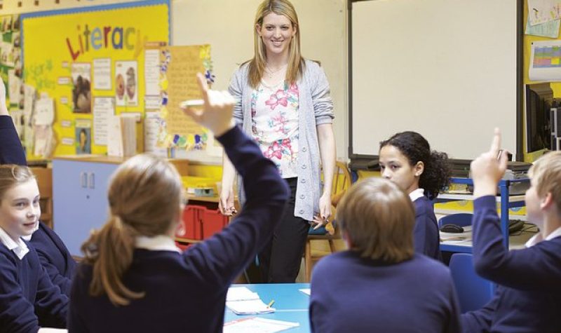 Une enseignante au cheveux blond est debout devant une classe. 5 élèves se retrouvent devant l'enseignante, dont une fille au centre et un gars à droite lèvent leur mains. À gauche, on peut apercevoir un tableau où il est inscrit «Literacy».