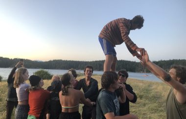 A teen boy stands on top of other teens shoulders and holds someone's hands for balance.