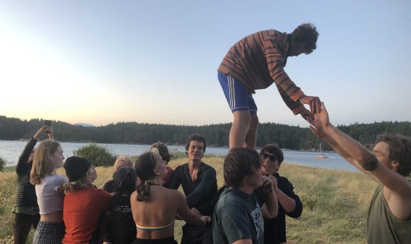 A teen boy stands on top of other teens shoulders and holds someone's hands for balance.