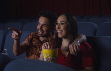 A man and a woman eating popcorn and watching a movie in a theatre. The seats are dark blue, set up in multiple rows.