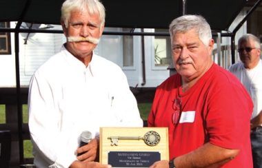 Terry Murdock, left, wearing a white collared shirt and sporting a large handlebar moustache hands an award to another man.