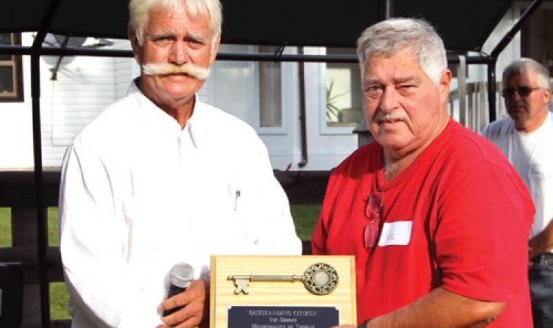 Terry Murdock, left, wearing a white collared shirt and sporting a large handlebar moustache hands an award to another man.