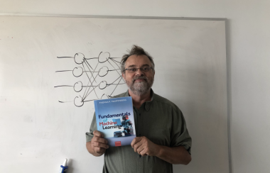 Le professeur devant un tableau représentant des réseaux neuronales et tenant son livre dans les mains.