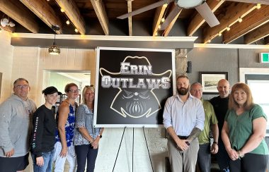 Off-ice staff for the Erin Outlaws stand by a sign of the team's logo, a bearded cowboy logo with the team's name.