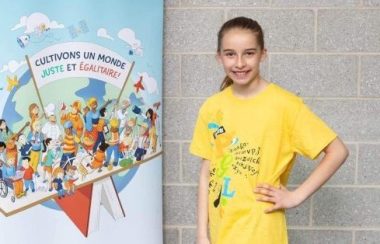 Une jeune fille portant un t-shirt jaune, pantalon noir et soulier noir à côté d'une affiche bleue à l'extérieur