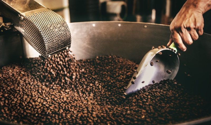 Coffee beans are coming down the chute into a metal pan where a hand stirs them with a scoop.