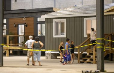People walking into an unfinished one story grey tiny homes behind yellow caution tape. There is also a dark brown home next to the home in the foreground.