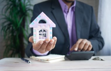 A man in a suit sits at a desk with a calculator, notebook and pen and holds up a model of a house.