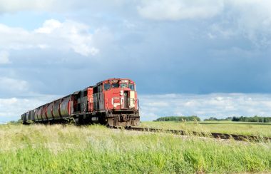 A red train pictures on traintracks travelling through a field on an overcast day.