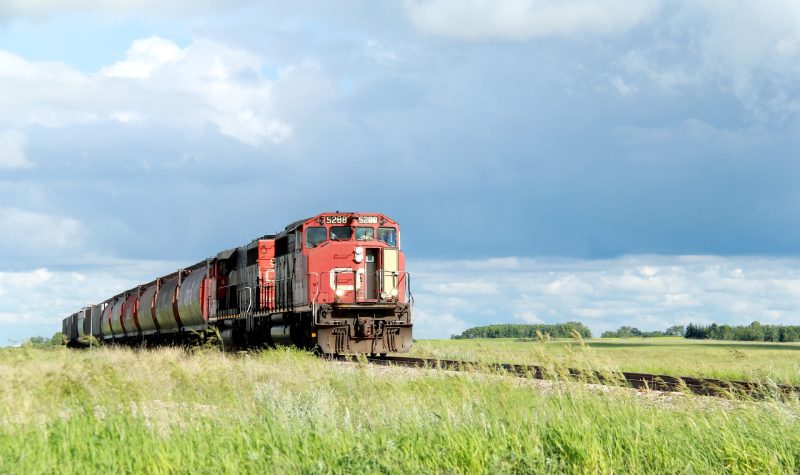 A red train pictures on traintracks travelling through a field on an overcast day.