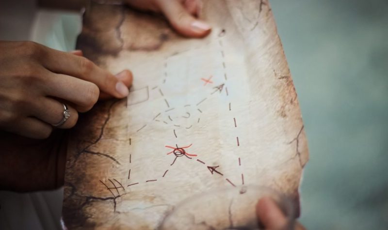 On aperçoit une carte aux trésor typique de l'univers des pirates. Des flèches noires et rouges sont tracés sur du papier jaunis et les contours sont abimés. On peut voir trois mains de personnes qui tienne la carte.