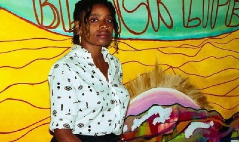 femme noire au chemisier blanc devant une oeuvre d'art : fond jaune avec écrit black life sur du vert
