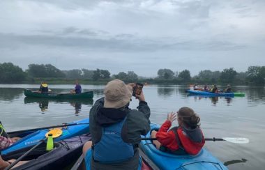 A group of people seated in canoes and kayaks on a river. One man is taking a picture of the landscape while others look on. Trees sit on each side of the river bank.