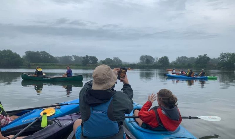 A group of people seated in canoes and kayaks on a river. One man is taking a picture of the landscape while others look on. Trees sit on each side of the river bank.