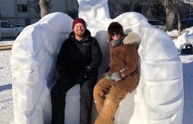 Nicolas et Françoise en manteau sont assit sur le banc de neige qui fait partie de la sculpture de hibou.