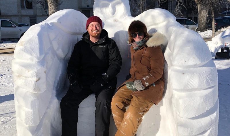 Nicolas et Françoise en manteau sont assit sur le banc de neige qui fait partie de la sculpture de hibou.