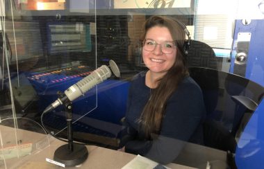 Josée est assise dans le studio de la radio devant un micro en portant des écouteurs. Elle porte un chandail bleu foncé à manche longue.