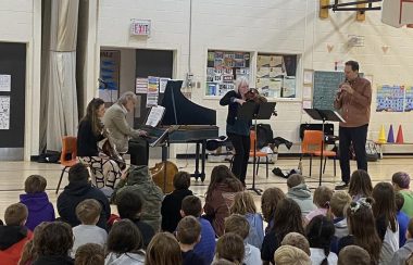 A group of school kids sitting on a gym floor looking at a group of four musicians playing a harpsichord, stringed instruments and a recorder.