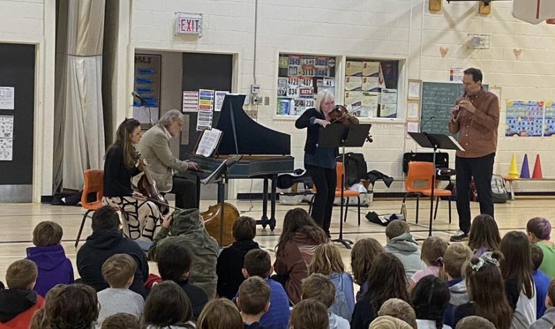 A group of school kids sitting on a gym floor looking at a group of four musicians playing a harpsichord, stringed instruments and a recorder.