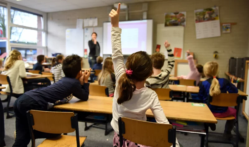 A child raises their hand in a busy classroom of youngsters at desks.