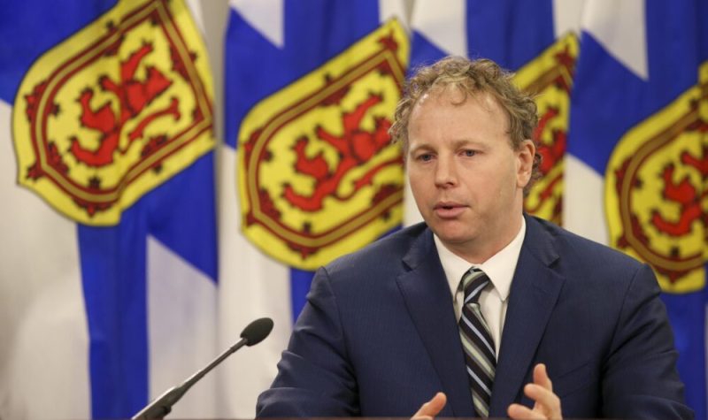 Homme avec gilet bleu, chemise blanche et cravate parlant dans ion micro avec des drapeaux de la Nouvelle-Écosse en arrière plan.