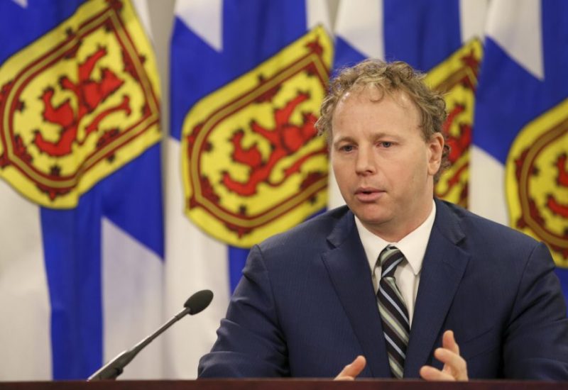 Homme avec gilet bleu, chemise blanche et cravate parlant dans ion micro avec des drapeaux de la Nouvelle-Écosse en arrière plan.