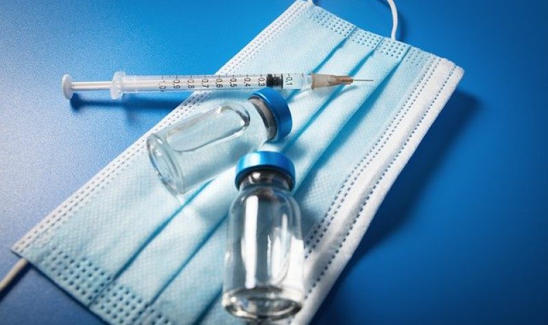 Un masque hygiénique sur une table bleue avec deux flacons de liquide vaccinal et une seringue.